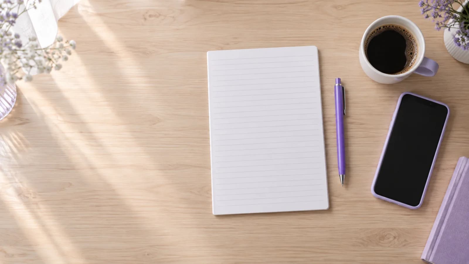 A wooden desk seen from above with a blank lined sheet of paper, a purple pen, a phone and a cup of coffee